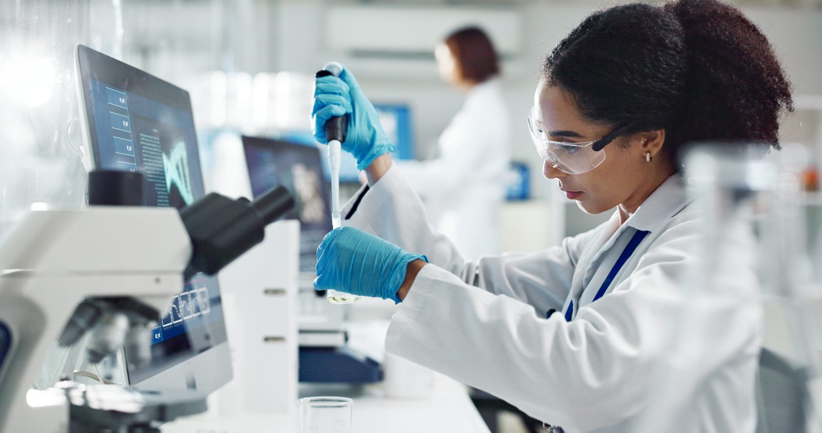 A scientist wearing safety glasses and blue gloves pipetting a sample at a lab bench.